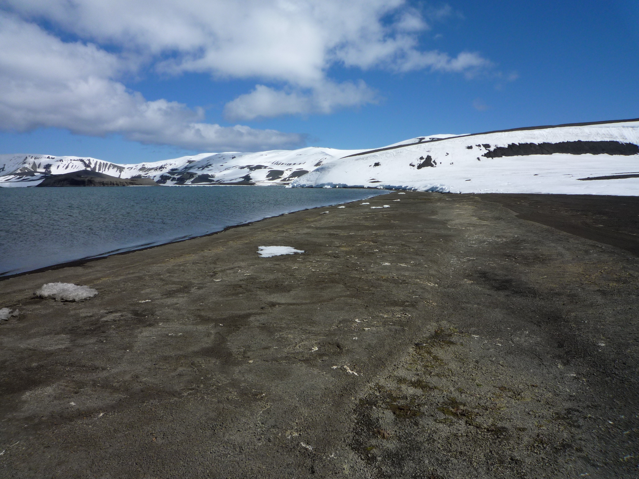 Zu sehen ist die Küste der Antarktis. Das Meer ist ruhig, der Himmel ist blau. Entlang der Küste erheben sich ein paar kleiner Hügel. Schnee und Eis sind zurückgegangen.