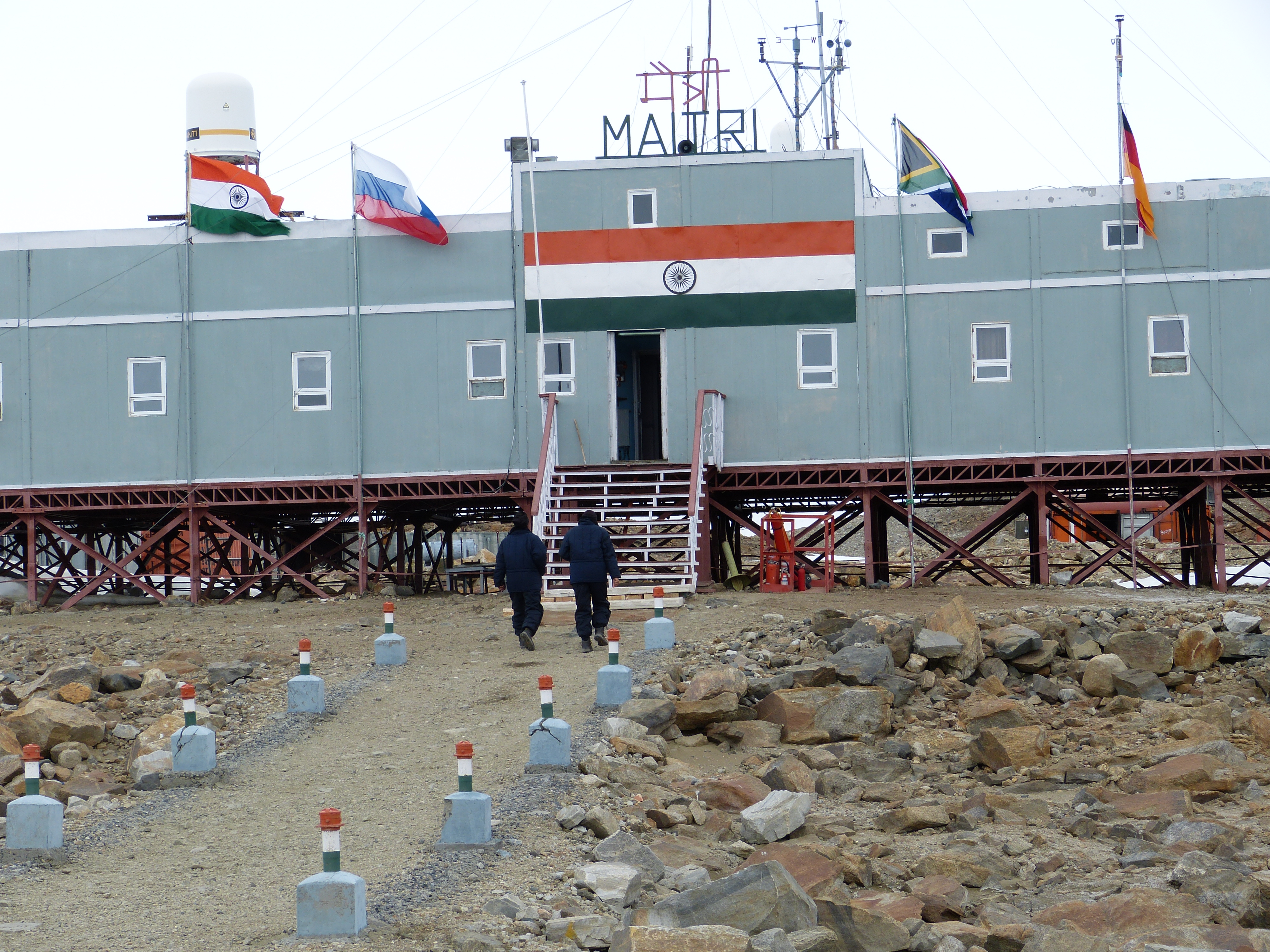 Im Bild ist die indische Forschungsstation auf deren Dach der Name der Station, "Maitri" steht. Davor stehen die Flaggen 4 anderer Länder. " Personen laufen auf die Station zu. 