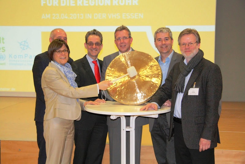 Hinter einem Stehtisch stehen sechs Personen. Sie posieren für ein Foto. Eine Frau hält eine große goldene Klangplatte und einen hölzernen Hammer,  mit dem sie auf die Platte schlägt, um einen Gong zu erzeugen. 