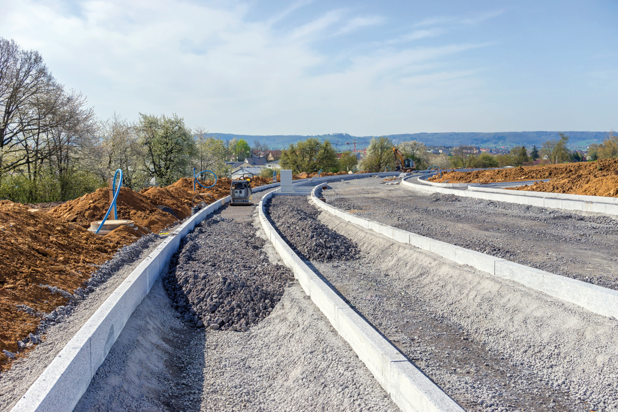 The picture shows the construction of a new road. The partly finished road foundation is visible. In the foreground the foundation is still being completed whereas in the background it is already sealed. Half-way along the site a vibratory plate is standing. To the left of the new road, mounds of earth are visible - behind those a treeline. In the background, the roofs of a residential area are visible.
