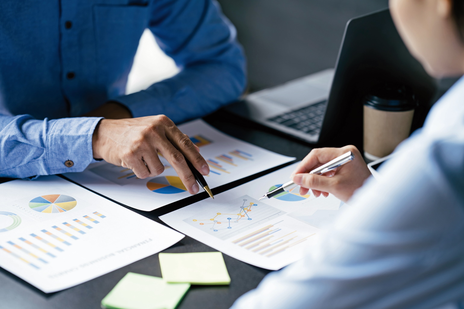 The picture shows the arms and hands of two formally dressed individuals sitting at a desk. There is a laptop on the desk, and there are documents which show various charts and sticky notes. Both individuals are holding ballpoint pens which they use to point to the same line chart.
