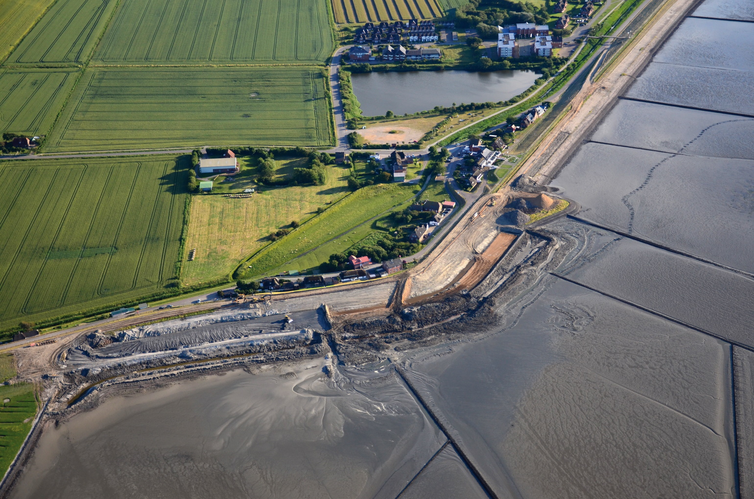 The picture shows an aerial view of a land protection dyke under construction. In the background, there are agricultural areas and houses as well as standing water visible.