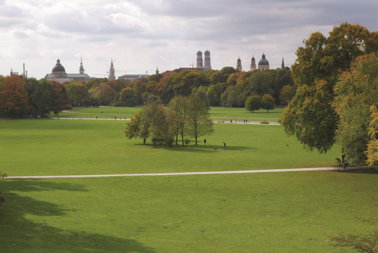 Das Bild zeigt den Blick in den Englischen Garten in München. Man sieht große Wiesenflächen, die von Wegen durchzogen sind und auf denen einzelne Baumgruppen stehen. Im Hintergrund sieht man die Stadtsilhouette von München. 