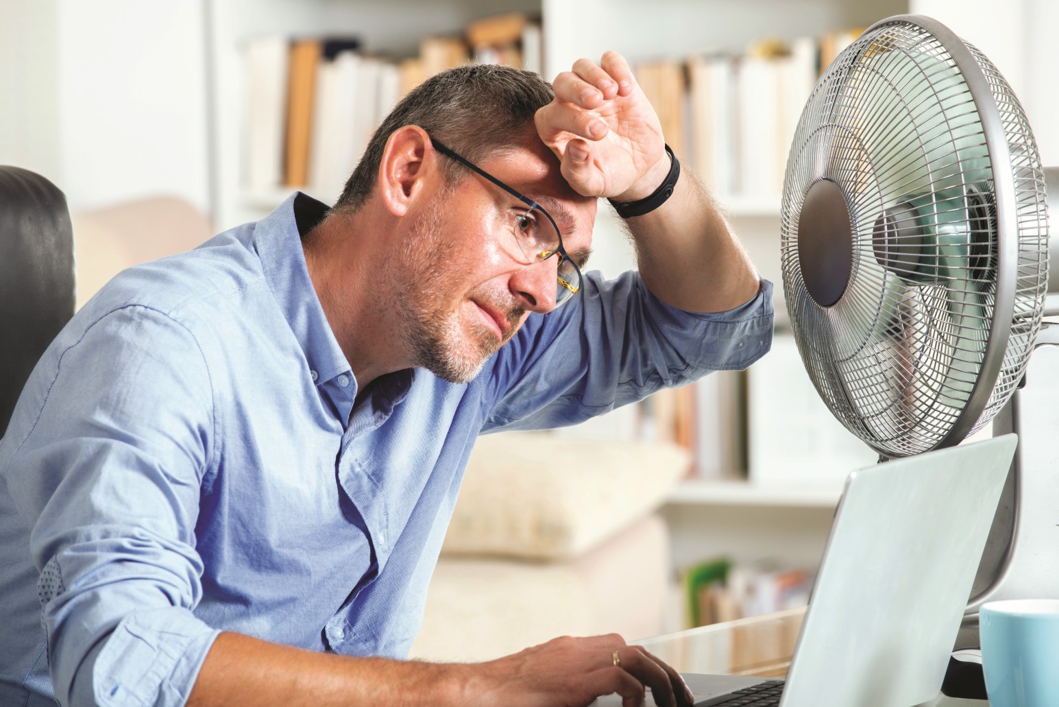The picture shows a man sitting in front of a fan in an office, holding the back of his hand to his forehead.