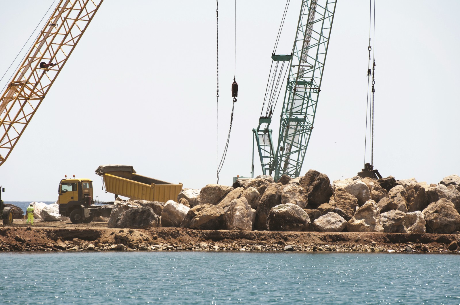 The picture shows a large construction project by the sea. Large blocks of stone are being moved by large cranes and a large truck. 