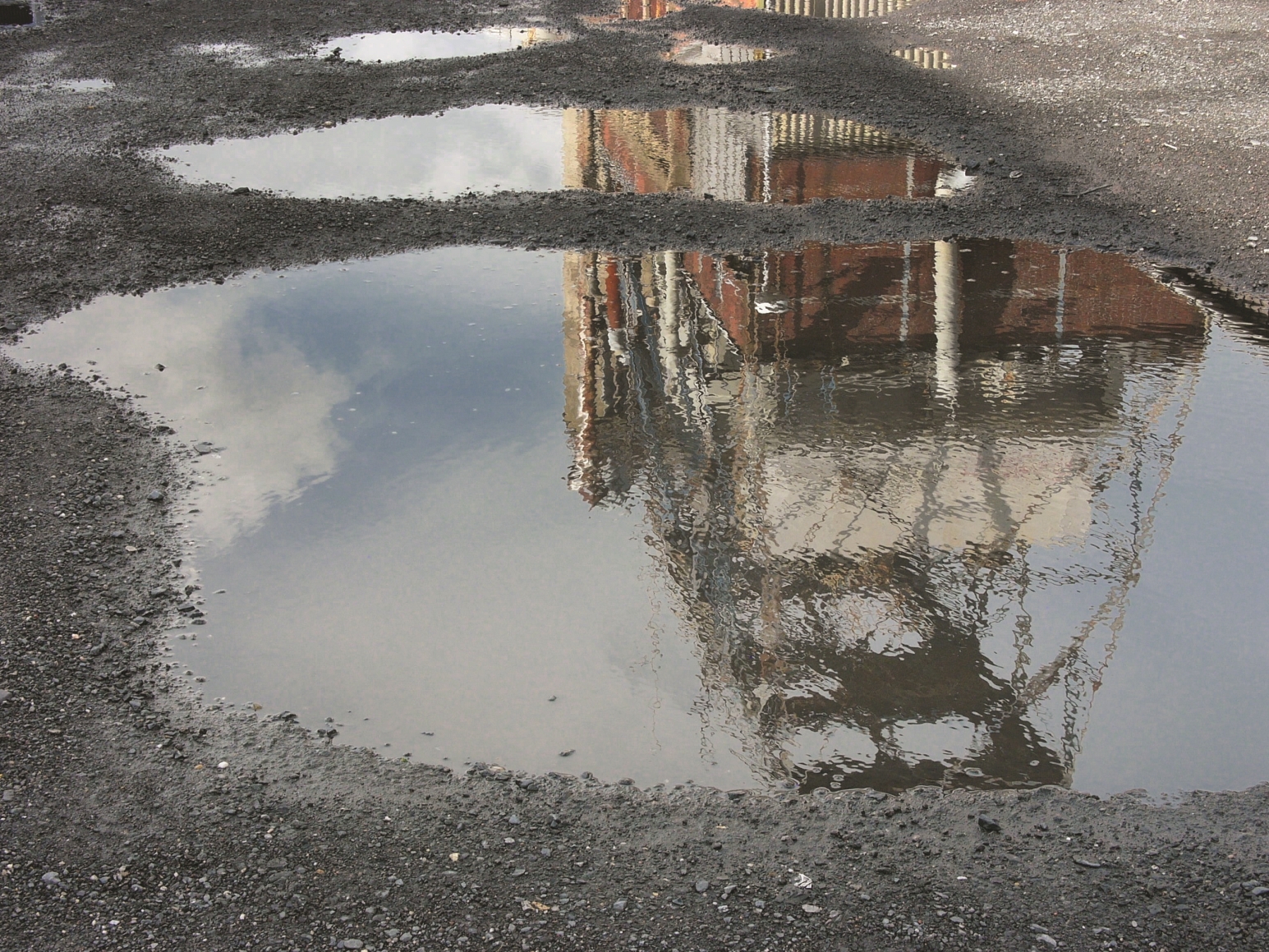 Das Bild zeigt zwei Pfützen auf einer Schotter-Kies-Fläche, in der sich ein Industriegebäude spiegelt.