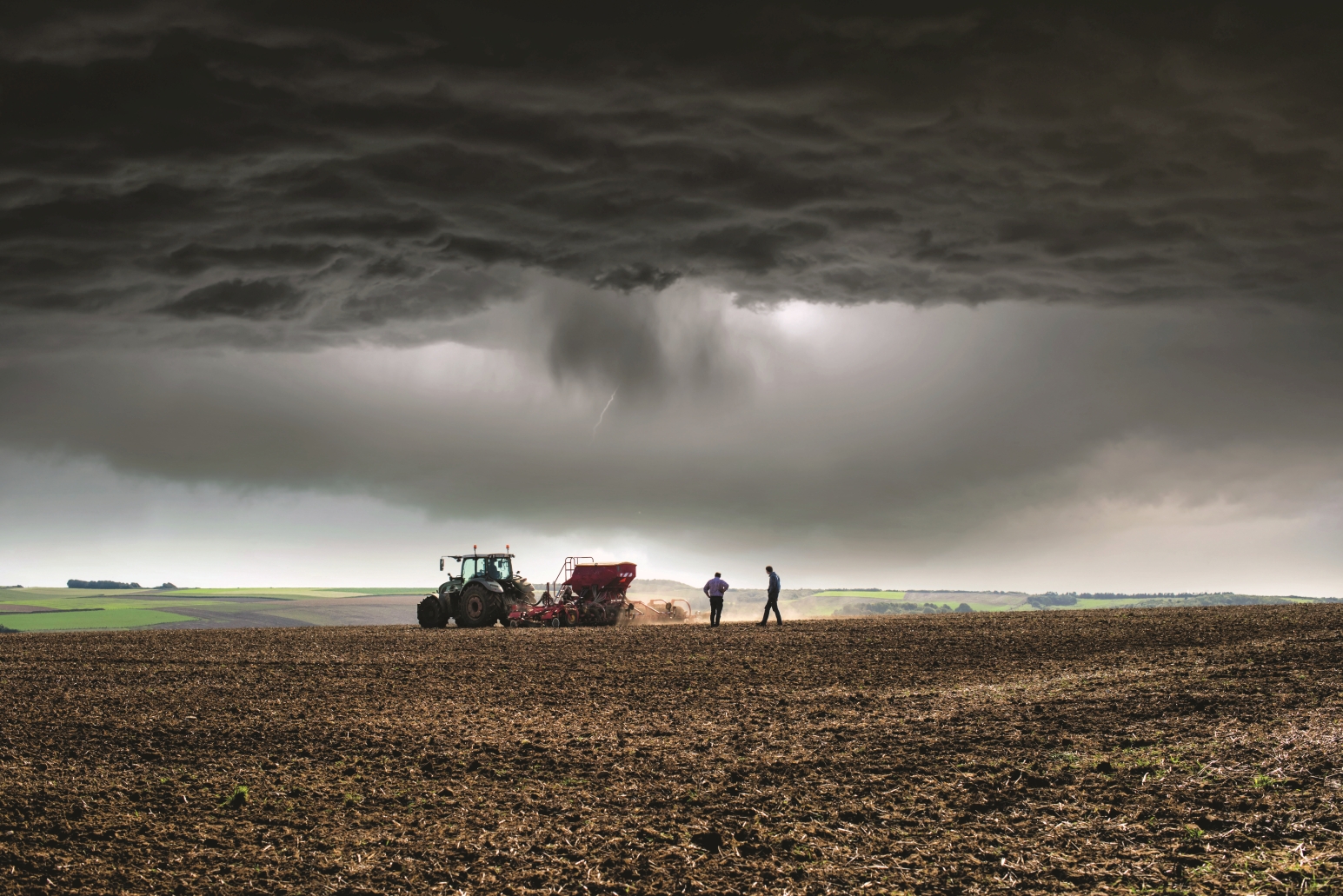 The picture shows a field in the foreground. A tractor is sowing and two men are checking the sowing. In the background, a wider agricultural landscape can be seen. The sky is very dark, a thunderstorm is approaching.