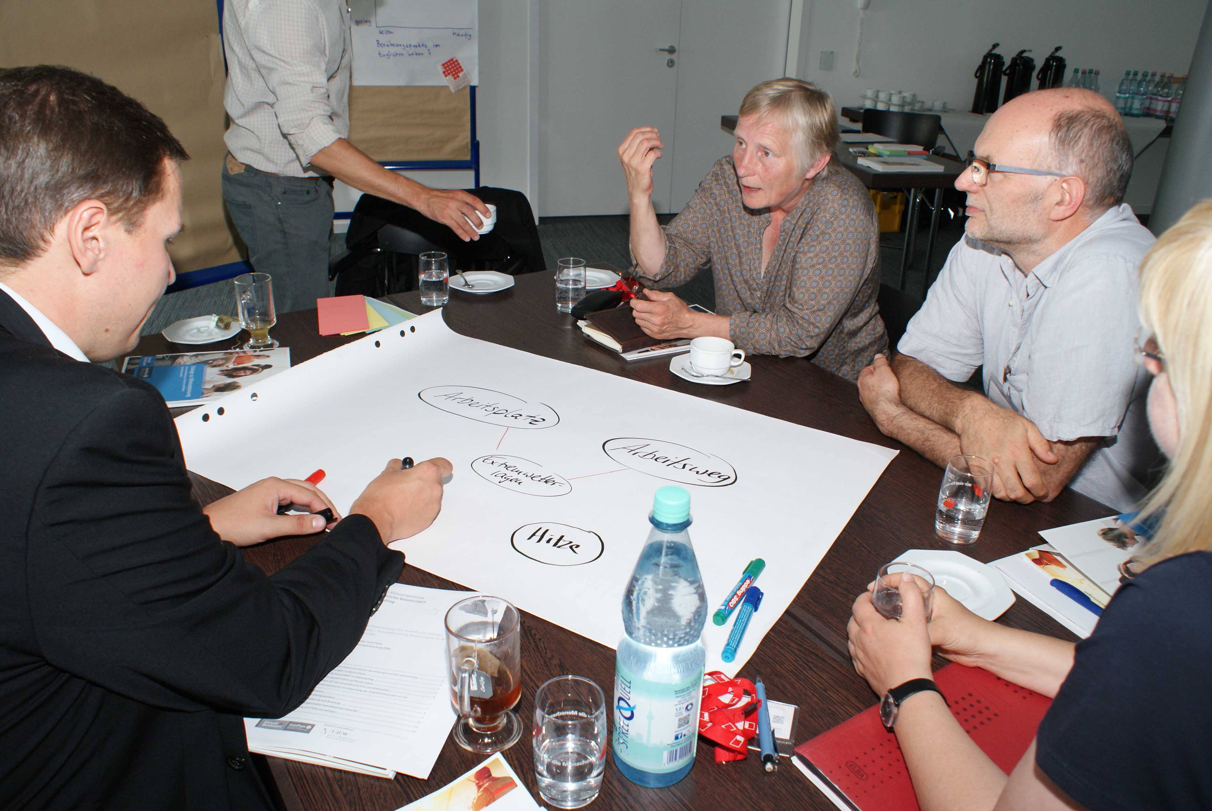 Teilnehmer sind am Gruppenarbeitstisch und diskutieren miteinander. Ein Teilnehmer schreibt Diskussionsergebnisse auf Posterpapier, welches auf dem Tisch liegt.