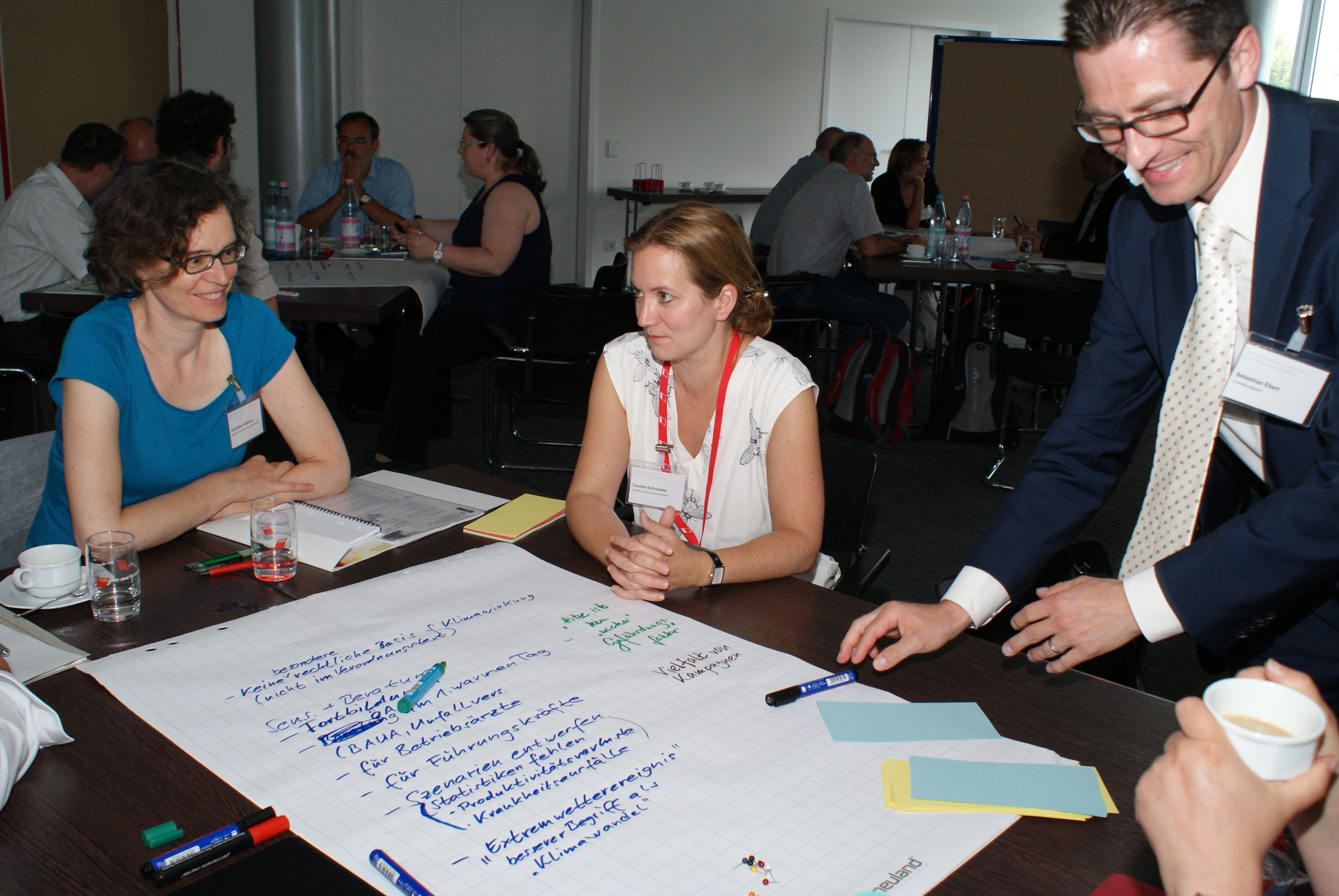 Teilnehmer sitzen am Gruppenarbeitstisch. Ein Teilnehmer steht am Tisch und legt den Stift aus der Hand, mit dem er die Diskussionsergebnisse der Diskutanten am Arbeitstisch auf das Posterpapier geschrieben hat. Die Teilnehmer schauen freundlich und lachen.