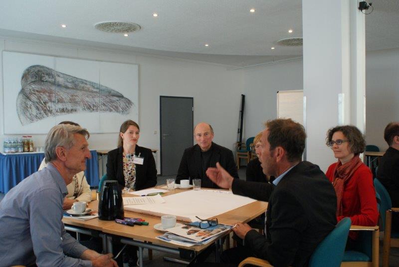 Teilnehmer sitzen diskutieren am Arbeitsgruppentisch und hören dem gestikulierenden Sprecher zu. Alle schauen freundlich drein.