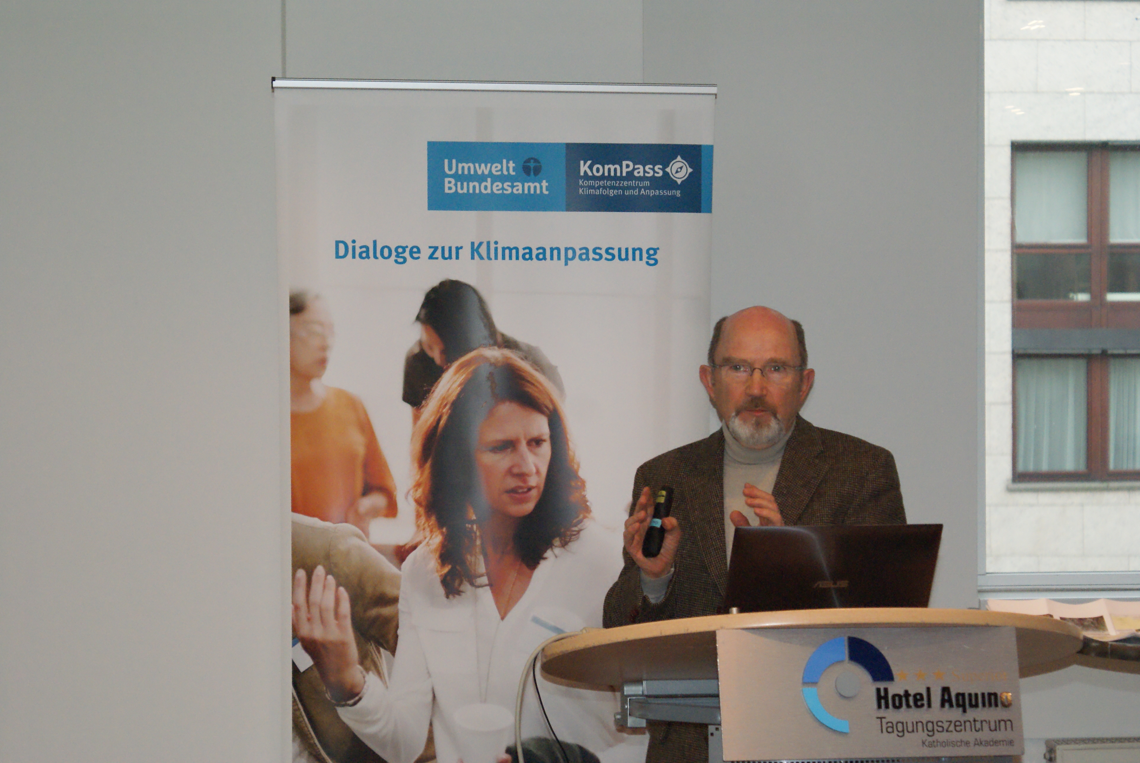 Vortragender steht am Rednerpult vor einem aufgeklappten Laptop und trägt gestikulierend vor. Vorne auf dem Stehpult steht der Name des Tagungszentrums Aquino und im Hintergrund steht das Rollup Dialoge der Klimaanpassung und man sieht rechts durch ein Fenster hinaus auf eine Häuserfassade.