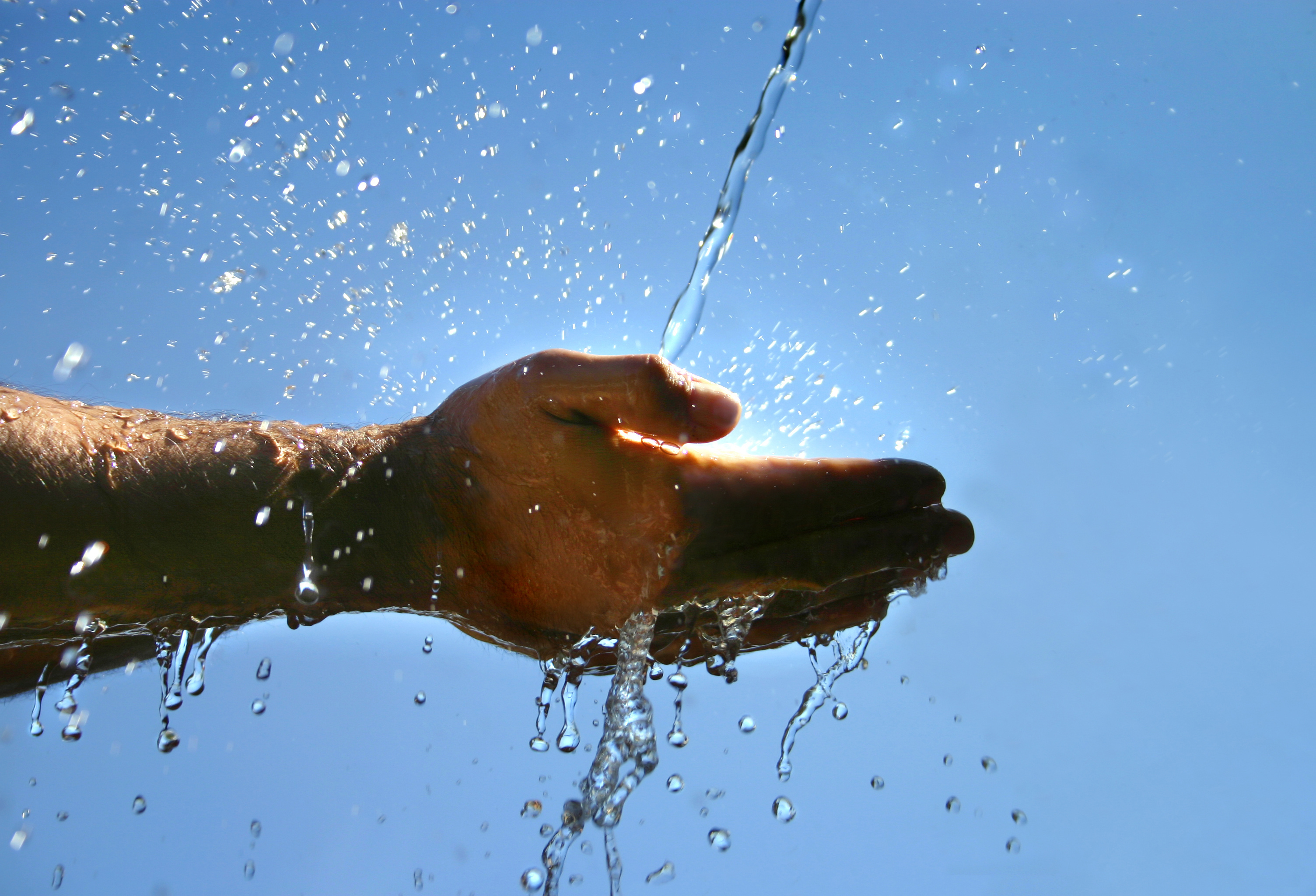 Zwei Hände werden in einen Wasserstrahl gehalten. Im Hintergrund scheint die Sonne