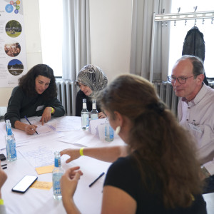 Das Foto zeigt einen Tisch mit mehreren Menschen, von denen eine Teilnehmerin Notizen macht.