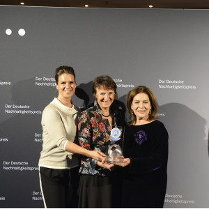 drei Frauen stehen lächelnd auf einer Bühne und halten eine "Blauer Engel"-Trophäe in die Kamera, im Hintergrund eine Wand mit Schriftzügen "Der Deutsche Nachhaltigkeitspreis"