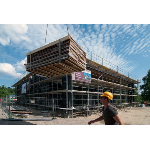 eingerüstetes Haus, davor ein Bauarbeiter mit gelbem Helm, ein Kran liefert Elemente für die Holz-Fassadenverkleidung an