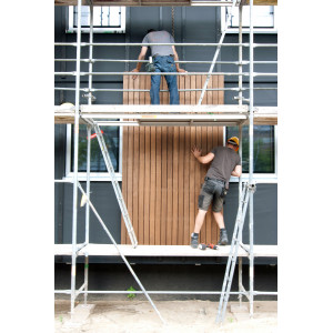 zwei Bauarbeiter stehen auf einem Baugerüst und montieren zusammen ein Holz-Fassadenelement