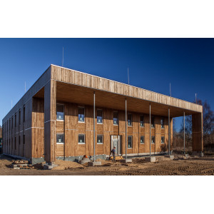 würfelförmiger, zweistöckiger Bau mit Holzfassade und Kollonadengang, davor ein Bauarbeiter, die Außenanlage noch ungestaltete Baustelle