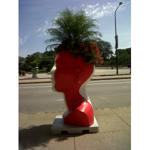 Auf dem Bürgersteig steht ein übergroßer roter Kopf, er dient als Blumentopf.