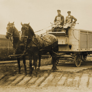 Pfedewagen mit zwei Arbeitern auf dem Kutschbock, die Ladefläche ist mit Metall-Rolläden abgedeckt. 