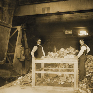 Zwei Arbeiter stehen zwischen einem Tisch, einem Berg Müll und einem Kran.