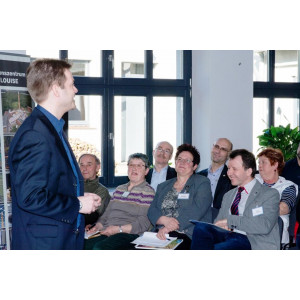 Ein Mann im Anzug steht vor einigen Personen im Publikum und spricht. Er hat die Hänge zusammen gefaltet und schaut geradeaus. Im Publikum sitzen 7 Personen in zwei Stuhlreihen hintereinander und hören zu. Sie tragen Namensschilder