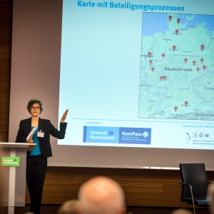 Vortragende steht am Rednerpult und zeigt auf Deutschlandkarte im Hintergrund. Die Karte zeigt an in Rot markiert, wo Beteiligungen bereits stattfinden.