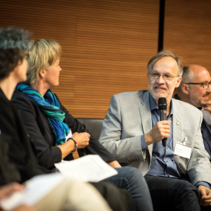 Personen sitzen zur Interviewrunde. Ein Teilnehmer hält das Mikrofon in der Hand und spricht hinein.