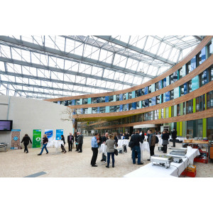 Veranstaltungsambiente im Forum des Umweltbundesamtes unter dem großzügigen Glasdach und vor der mit Holz verkleideten Fassade mit viel Fensterglas in den Farben Grün und Blau abgesetzt. Auf der Steinfläche am Boden stehen die Teilnehmenden an Stehtischen und diskutieren.