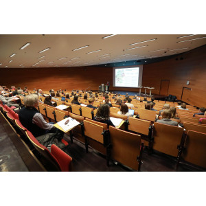 Veranstaltungsambiente im Hörsaal des Umweltbundesamtes. Der Blick geht nach vorn Richtung Podium zur Projektionsfläche. Ein Teilnehmener steht auf dem Podium am Stehpult und präsentiert seinen Vortrag. Die Teilnehmenden sitzen davor und hören zu. Der Hörsaal ist in warmen Holzfarben gehalten.
