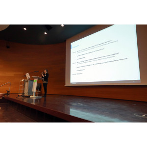 Vortragende steht auf dem Podium an einem Rednerpult und trägt vor mit einem Mikrofon in der Hand. Im Hintergrund ist das Veranstaltungsprogramm auf der Projektionsfläche zu sehen.