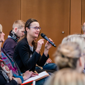 Eine Teilnehmerin spricht angeregt ins Handmikrofon. Alle Teilnehmende sitzen in Reihen im Veranstaltungsraum. Die Wände sind holzvertäfelt.