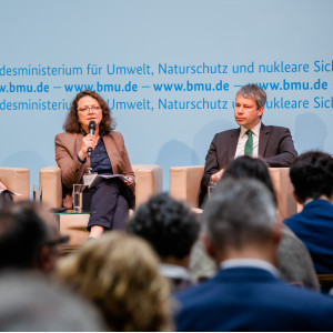 Podiumsdiskussion vor hellblauem Hintergrund, auf dem der Veranstaltungsort geschrieben steht.