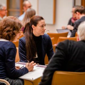 Teilnehmende haben sich in Gruppen im Veranstaltungsraum zusammengesetzt auf orangenen Stühlen und sprechen miteinander. Ergebnisse werden dabei auf Arbeitsbögen von einer Teilnehmerin skizziert.