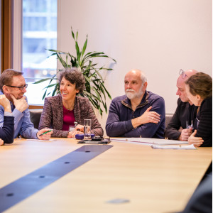 Teilnehmende sitzen an einem großen hellfarbenen Arbeitstisch und tauschen sich lachend miteinander aus. Im Hintergrund geht der Blick durch ein Fenster an einer Grünpflanze hinaus auf ein Gebäudeteil.
