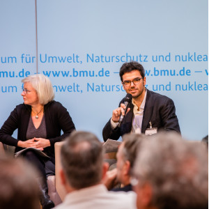 Podiumsdiskussion vor dem hellblauem Hintergrund, auf dem der Veranstaltungsort geschrieben steht. Ein Teilnehmender hält ein Handmikrofon.