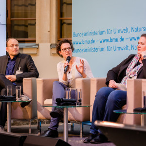 Podiumsdiskussion vor dem hellblauem Hintergrund, auf dem der Veranstaltungsort geschrieben steht. Eine Teilnehmende hält ein Handmikrofon und spricht angeregt zu den anderen Podiumstteilnehmenden.