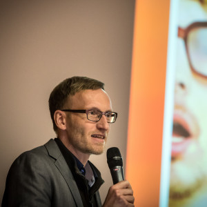 Vortragender steht mit Mikrofon in der Hand neben seiner Präsentation.