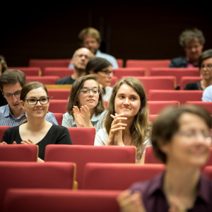 Teilnehmerinnen und Teilnehmer sitzen applaudierend in den roten Stuhlreihen des Hörsaales.