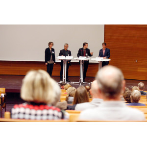 Teilnehmende diskutieren miteinander auf dem Podium. Sie stehen nebeneinander an Stehtischen aufgereiht. Der blick geht von hinten zwischen den Zuhörenden durch nach vorne.