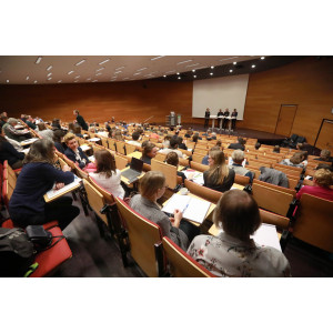 Veranstaltungsambiente im Hörsaal des Umweltbundesamtes: Der Blick geht nach vorne über die sitzenden Teilnehmenden hinweg zum Podium hin. Auf dem Podium stehen Diskutanten und Diskutantinnen an Stehtischen.