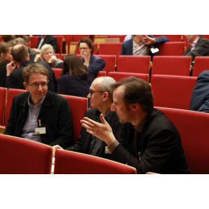 Teilnehmende sitzen auf den roten Sitzen im Hörsaal und sprechen miteinander. Ein Teilnehmer hebt dabei gestikulierend seine linke Hand.