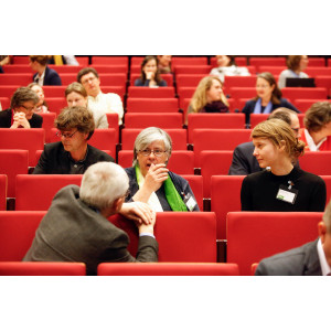 Im Hörsaal sitzen die Teilnehmenden auf den roten Sitzen und diskutieren miteinander. Eine Teilnehmende mit grünem Tuch um dem Hals gestikuliert dabei mit der rechten Hand.