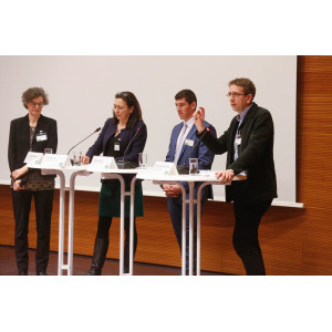 Teilnehmende stehen an Stehtischen auf dem Podium. Auf den Stehtischen stehen Gläser mit Wasser. Namensschilder und Tischmikrofone. Ein Teilnehmer spricht gestikulierend ins Publikum.