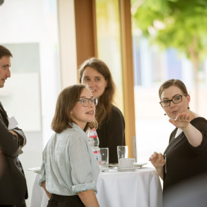 Teilnehmer und Teilnehmerinnen stehen in der Pause am Bistrotisch. Sie folgen schauend dem Hinweis einer Teilnehmerin, die in eine Richtung zeigt. Auf dem Bistrotisch stehen Gläser, Tassen und Teller sowie ein Getränkeflasche.Auf dem B