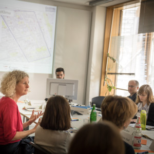 Workshopgruppen Teilnehmerinnen und Teilnehmer sitzen am Tisch und hören Teilnehmerin im roten Pullover zu. Im Hintergrund ist die Präsentation von Kiez Klima zu sehen und ein Teilnehmer sitzt hinter einem Bildschirm.