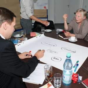 Teilnehmer sind am Gruppenarbeitstisch und diskutieren miteinander. Ein Teilnehmer schreibt Diskussionsergebnisse auf Posterpapier, welches auf dem Tisch liegt.