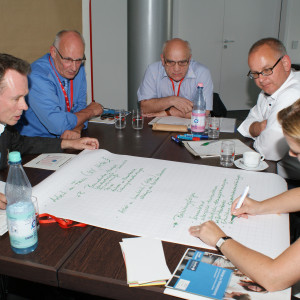 Teilnehmer sitzen im Arbeitsgruppenraum am Arbeitsgruppentisch und schauen auf das, was eine Teilnehmerin auf dem auf dem Tisch liegendes große Blatt Papier schreibt.
