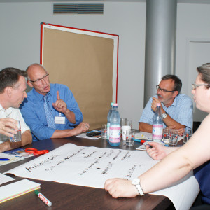 Teilnehmer diskutieren am Gruppenarbeitstisch. Eine Teilnehmerin schreibt Diskussionsergebnisse auf Posterpapier, welches auf dem Tisch liegt. Erfrischungsgetränke stehen auf dem Tisch. Ein Teilnehmer trinkt aus einem Glas Mineralwasser.