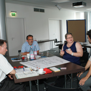 Teilnehmer sitzen diskutierend am Gruppenarbeitstisch. Auf dem Tisch liegt das beschriebene Posterpapier mit den Diskussionsergebnissen. Im Hintergrund stehen andere diskutierende Teilnehmer an einer Moderationswand.