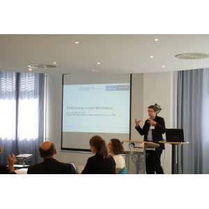Ein Vortragender steht am Rednerpult und eröffnet gestikulierend den Dialog. Im Hintergrund erscheint das Eröffnungbild des Dialogs mit Titel und Logo.